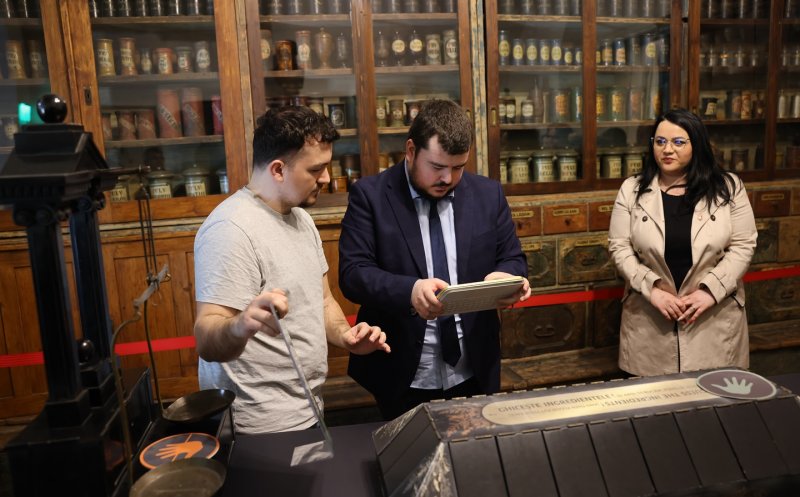 Trei persoane într-un spațiu muzeal sau expozițional, în fața unui stand interactiv. În stânga, un bărbat ține în mână un obiect metalic și pare să explice sau să demonstreze ceva. În centru, un alt bărbat îmbrăcat în costum privește atent un dispozitiv sau o tabletă pe care o ține în mâini. În dreapta, o femeie stă și observă activitatea. În fundal se află dulapuri din lemn cu rafturi pline de recipiente și borcane vechi, sugerând un context istoric sau științific. În prim-plan se vede un panou interactiv cu simboluri tactile, indicând că expoziția este gândită și pentru accesibilitate.