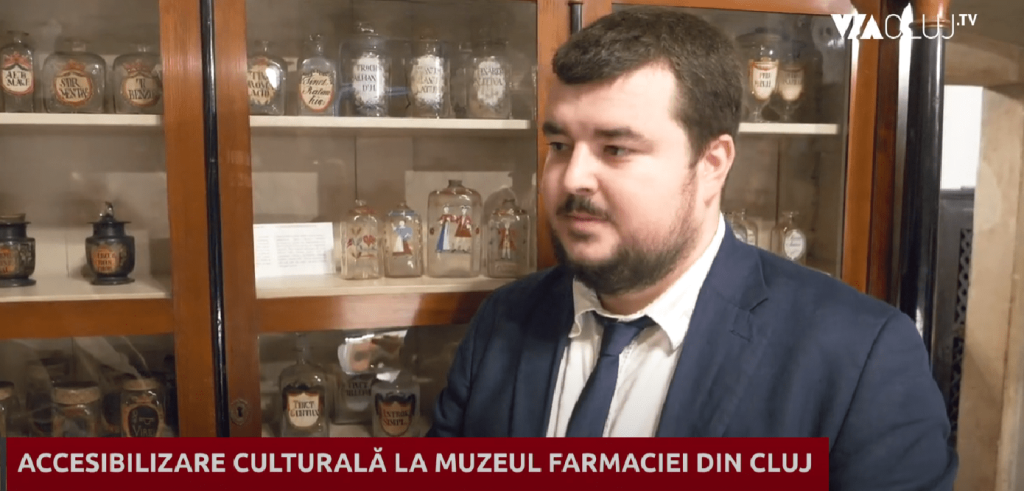 Un bărbat îmbrăcat în costum și cravată este surprins într-un interviu, într-un spațiu muzeal. În fundal se află un dulap din lemn cu uși din sticlă, plin cu recipiente și borcane vechi, specifice unui muzeu al farmaciei. În partea de jos a imaginii apare un banner roșu cu textul „ACCESIBILIZARE CULTURALĂ LA MUZEUL FARMACIEI DIN CLUJ”, iar în colțul din dreapta sus este vizibil logo-ul Cluj TV.