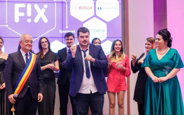 Un grup de persoane se află pe o scenă, în cadrul unui eveniment. În centru, un bărbat îmbrăcat în costum face un gest expresiv cu mâinile, fiind în prim-plan. În jurul lui sunt mai multe persoane îmbrăcate elegant, care zâmbesc și participă la moment. În stânga se află un bărbat care poartă eșarfă tricoloră, sugerând o funcție oficială. În fundal se vede un ecran mare cu logo-ul „FIX” și numele unor sponsori, indicând un eveniment formal sau o gală. Atmosfera este festivă și dinamică.